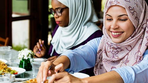 Two Muslim women in traditional attire dining in a worksite cafeteria.