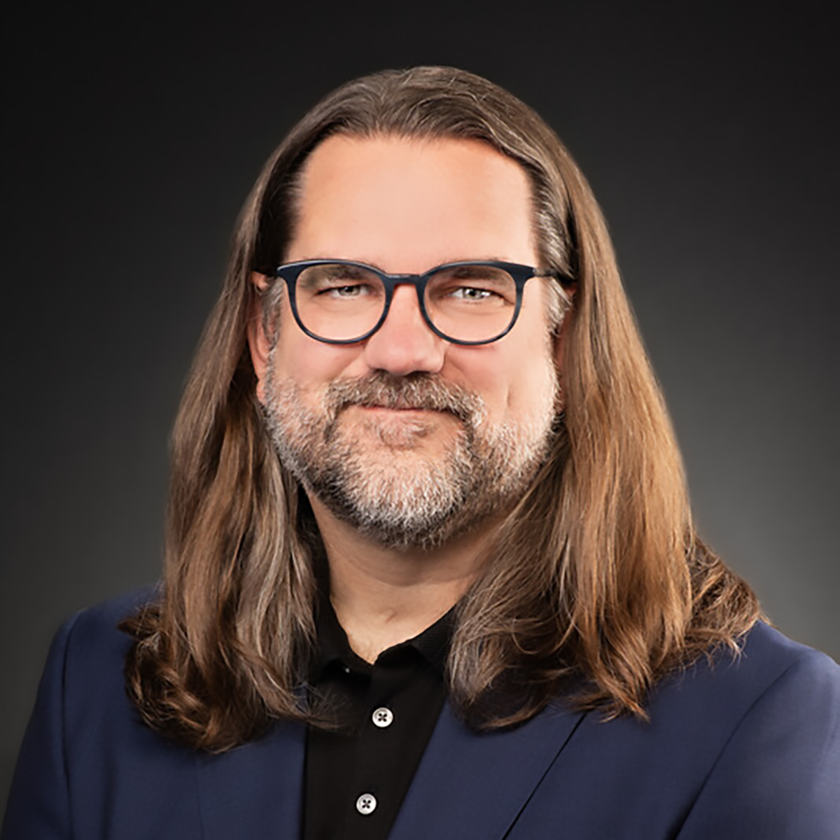 Dr. Dylan George, CFA's director, with long brown hair and short brown beard, wearing a dark blue shirt against a grey background.