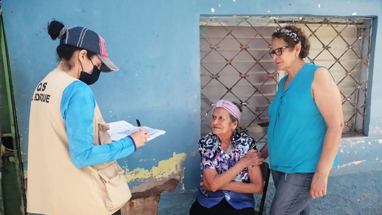 — title missing — A masked health worker holds a notebook and speaks with two local women about vaccinations in Honduras