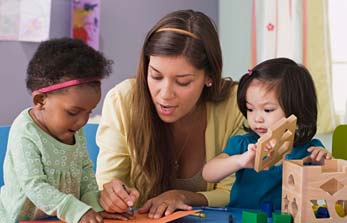 A teacher is helping a toddler draw while another toddler is stacking blocks.