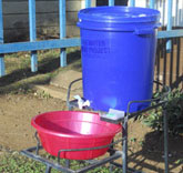 Handwashing station with plastic bucket containing a tap, metal stand, basin for catching water and soap.