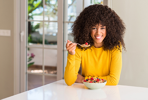 African american woman eating cereals, raspberries and blueberries with a happy face standing and smiling with a confident smile showing teeth