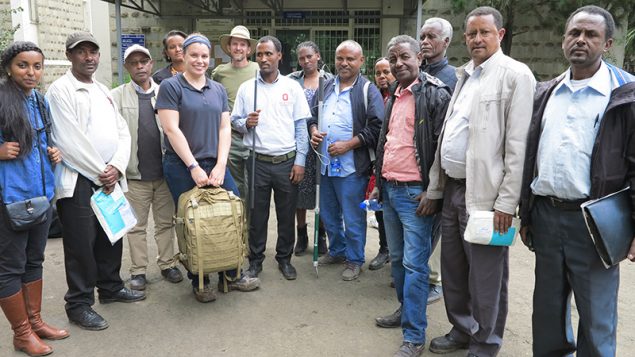 14 people standing in a half circle facing the camera, most with papers, packages, or other gear for their work