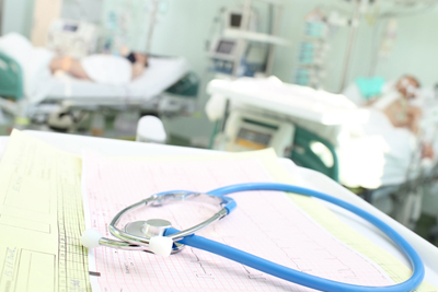 An image with a stethoscope laying on paper in the forefront with people lying in hospital beds in background.