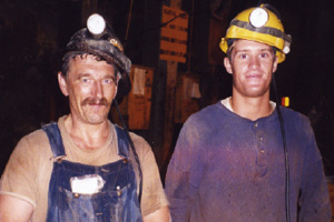 Two men look directly at the camera and grin. They are both very dirty. One man in overalls and t-shirt wearing a black miner&rsquo;s hardhat with a light attached to the front looks to be about 50. Standing very close to this man is a younger man, probably in his twenties. He is wearing a dirty blue shirt and a yellow miner&rsquo;s hard hat with a light attached to the front and a smaller one just above his right ear.