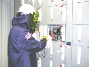 An electrician using a meter to test a 480V circuit, while wearing electrically-rated gloves, arc-rated face shield, and an arc-rated protective jacket.