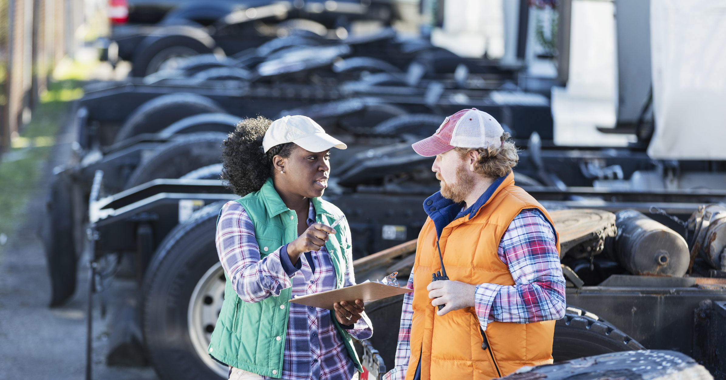 Multi-ethnic workers at a trucking company, standing by a fleet of semi-trucks, conversing.