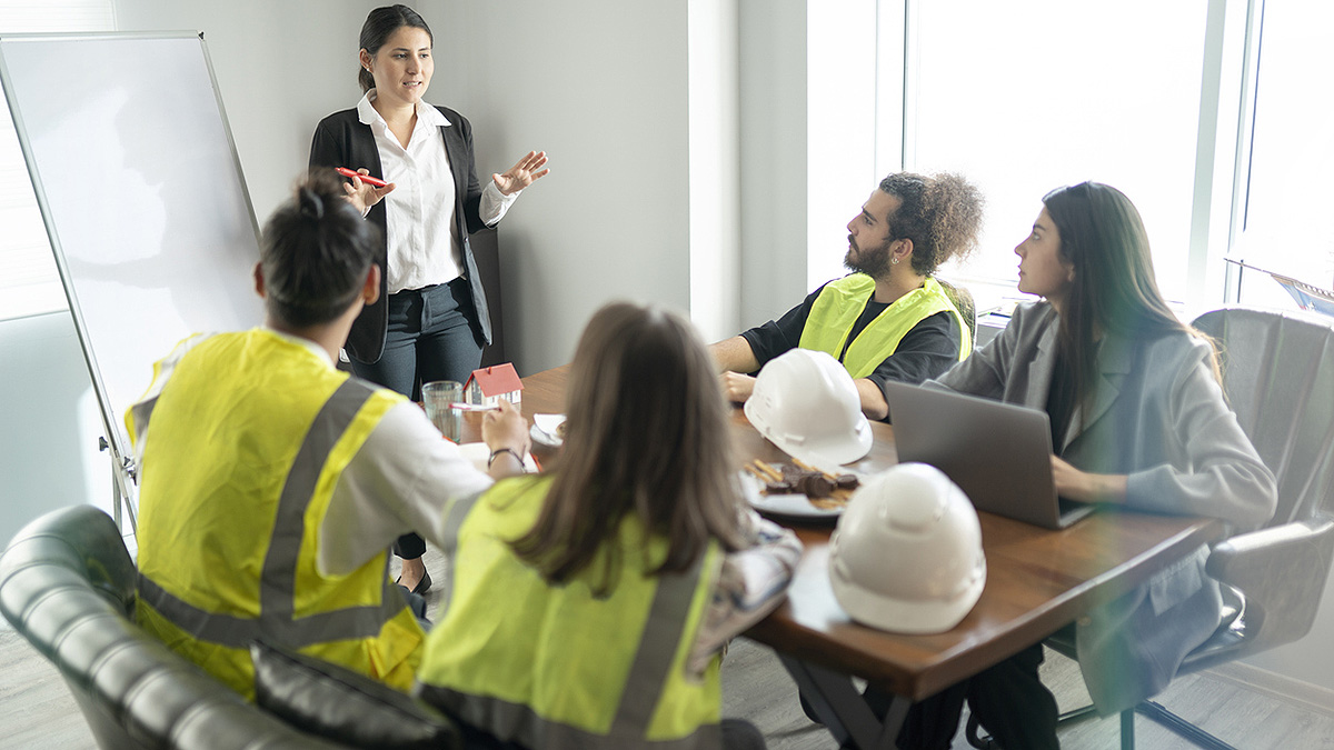Businesswoman standing at dry erase board training three workers in hardhat gear and one woman in business attire.